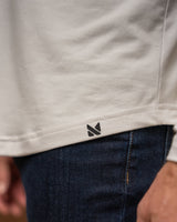 Close-up of a person wearing a light beige or off-white t-shirt and dark blue jeans. The t-shirt has a small black geometric logo near the hem.
