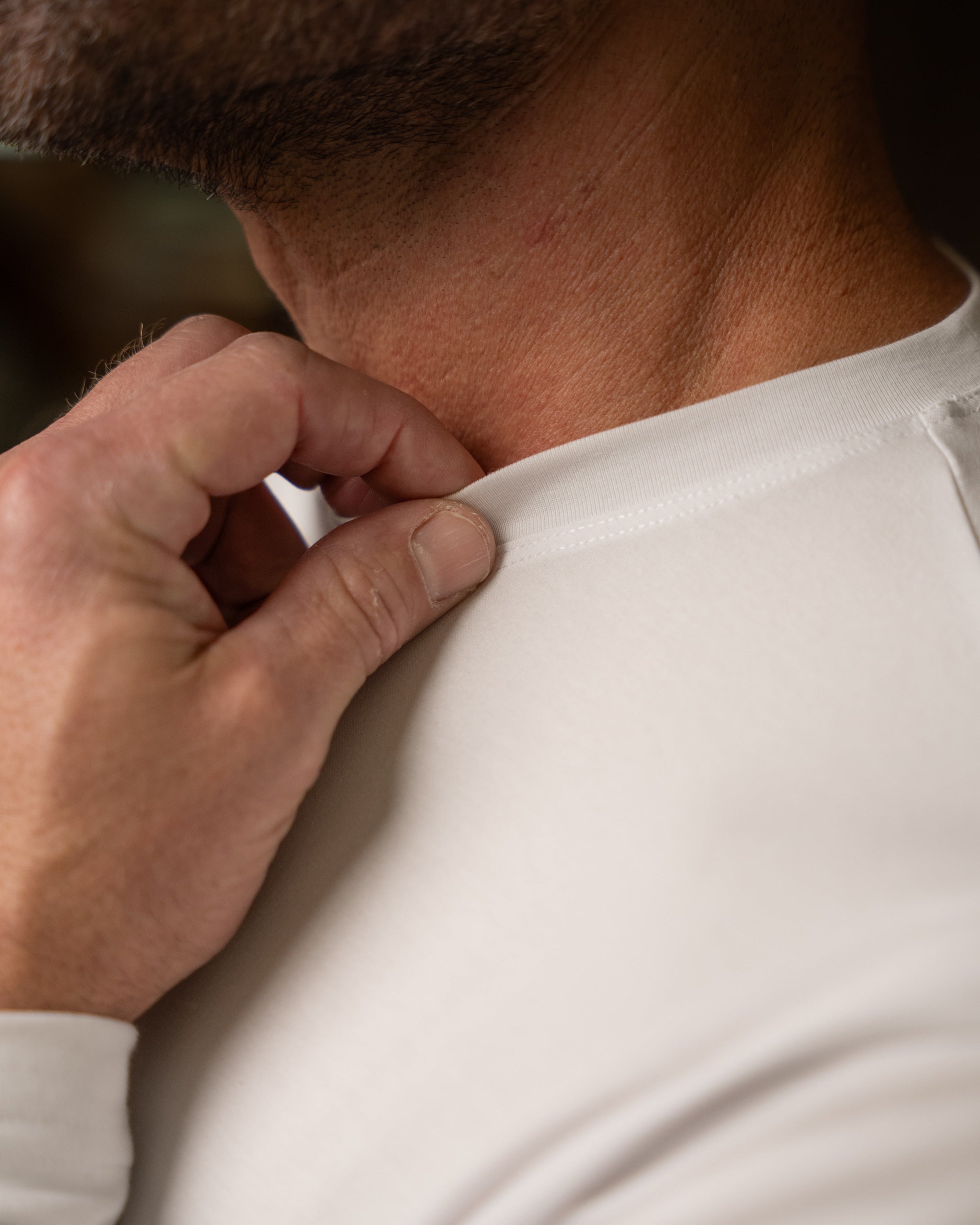 Close-up of a man's neck and shoulder wearing a white long-sleeve shirt, with his hand pulling the collar slightly outward to demonstrate stretch.