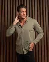 A man wearing a long-sleeve button-up shirt in a muted olive green color, posed against a dark brown vertical slat wall.