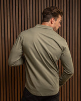 A man wearing a long-sleeve olive green shirt and black pants, shown from behind against a dark brown vertical slat wall.