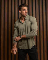 A man wearing a long-sleeve olive green button-down shirt and black pants, standing against a dark brown wood slat wall.