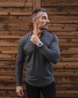 A man wearing a dark gray long-sleeve athletic shirt and black pants, standing against a rustic wooden plank wall and looking off-camera while wearing a watch.