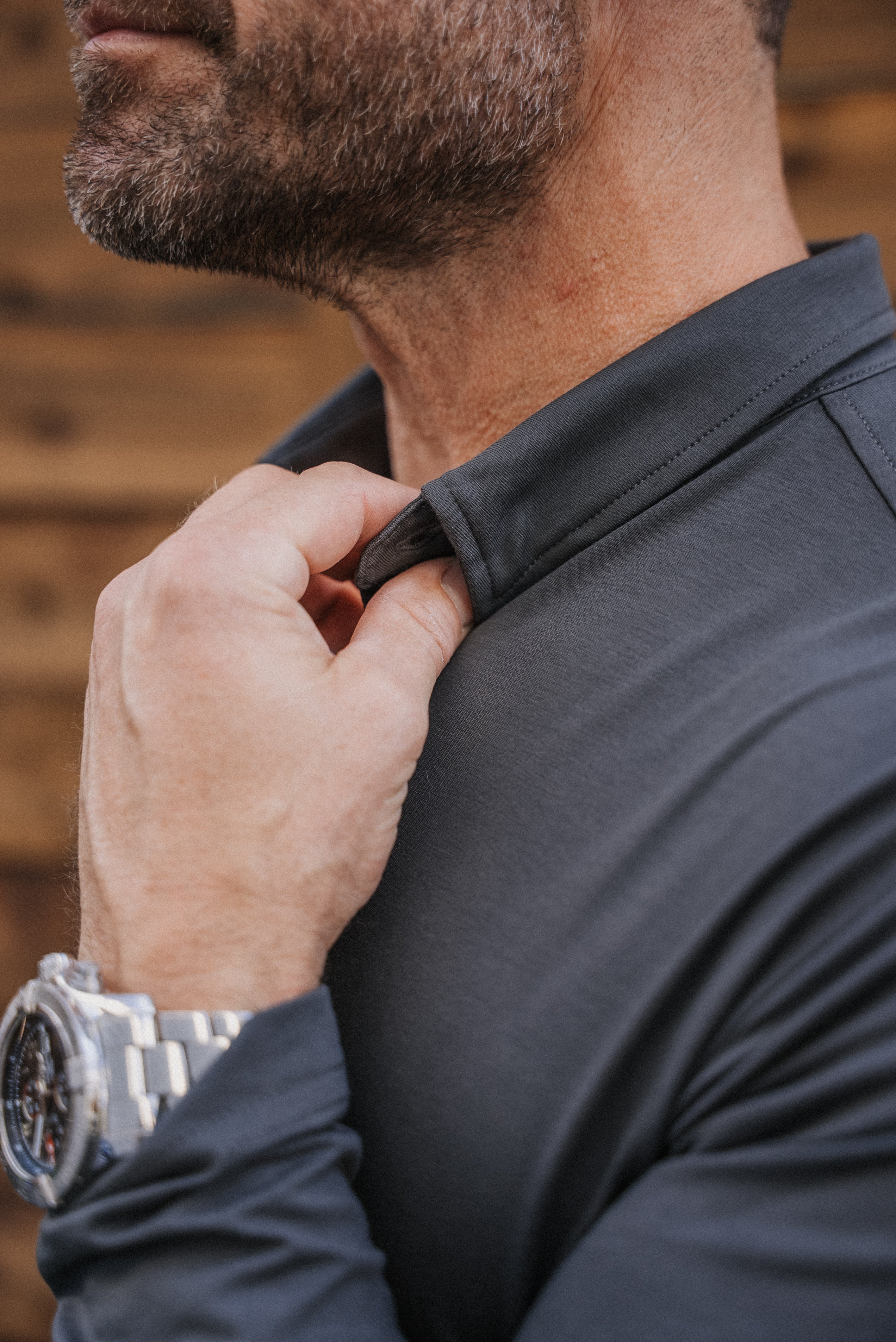 Close-up of a man wearing a dark charcoal gray long-sleeve performance shirt, adjusting the collar with one hand. He is wearing a silver metal watch on his wrist, and the background is a warm, blurred wood panel wall.