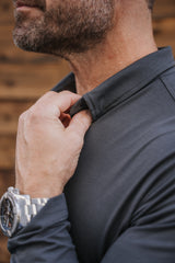 Close-up of a man wearing a dark charcoal gray long-sleeve performance shirt, adjusting the collar with one hand. He is wearing a silver metal watch on his wrist, and the background is a warm, blurred wood panel wall.