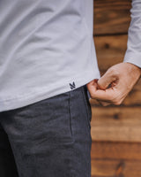Close-up of a person wearing a white long-sleeve shirt and dark charcoal pants, pulling the hem of the shirt to reveal a small black embroidered logo, with a wooden wall background.