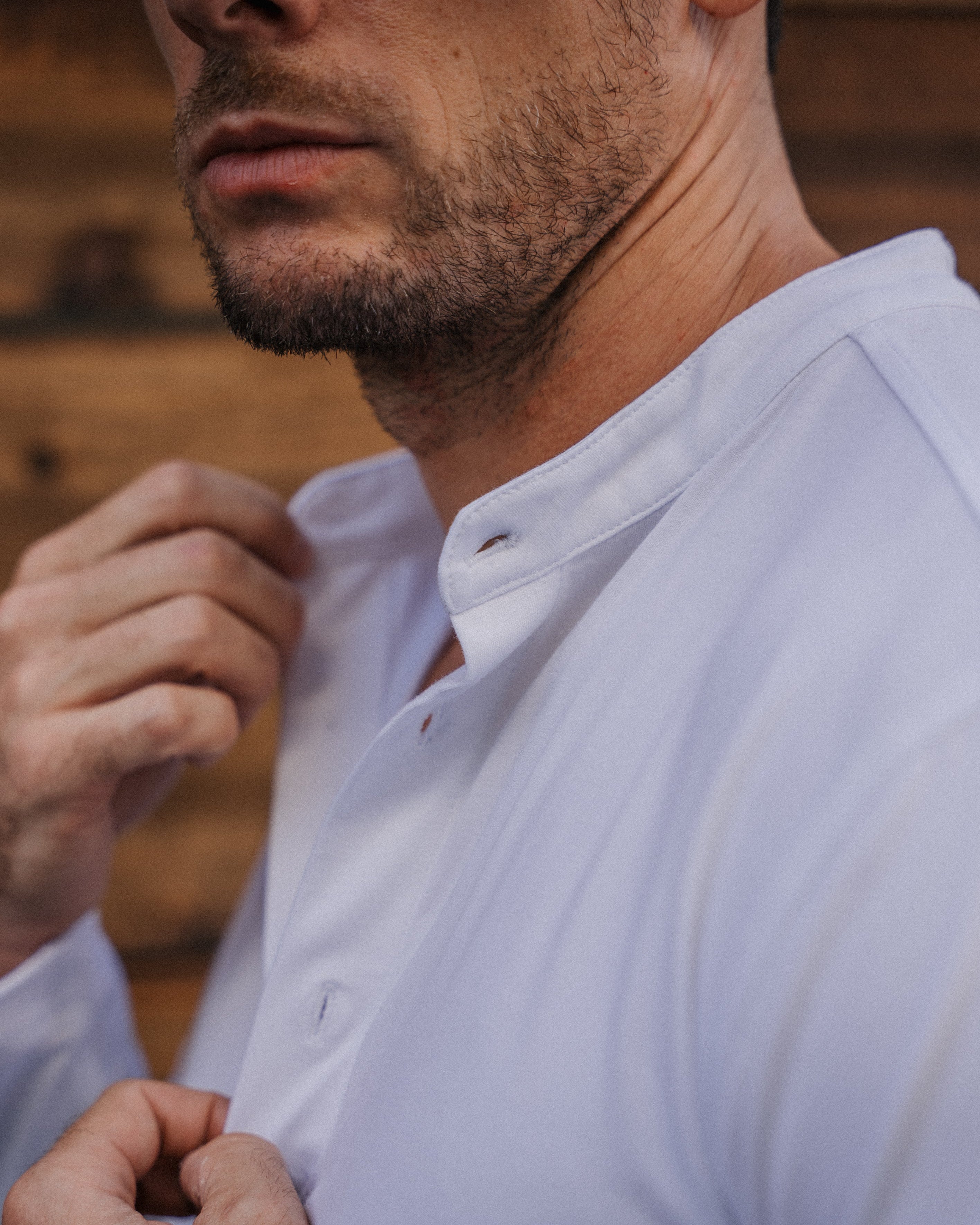 Close-up of a man wearing a white long-sleeve button-up shirt with a mandarin collar, adjusting the front buttons. The background is a rustic wooden wall.