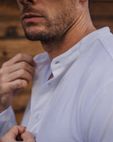 Close-up of a man wearing a white long-sleeve button-up shirt with a mandarin collar, adjusting the front buttons. The background is a rustic wooden wall.