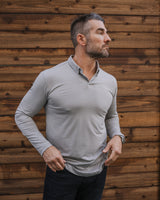 A man wearing a light gray long-sleeve polo shirt and dark jeans, standing against a rustic brown wooden plank wall.
