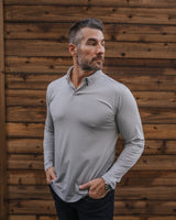 A man wearing a light gray long-sleeve collared performance shirt and dark pants, standing against a rustic brown wooden plank wall.