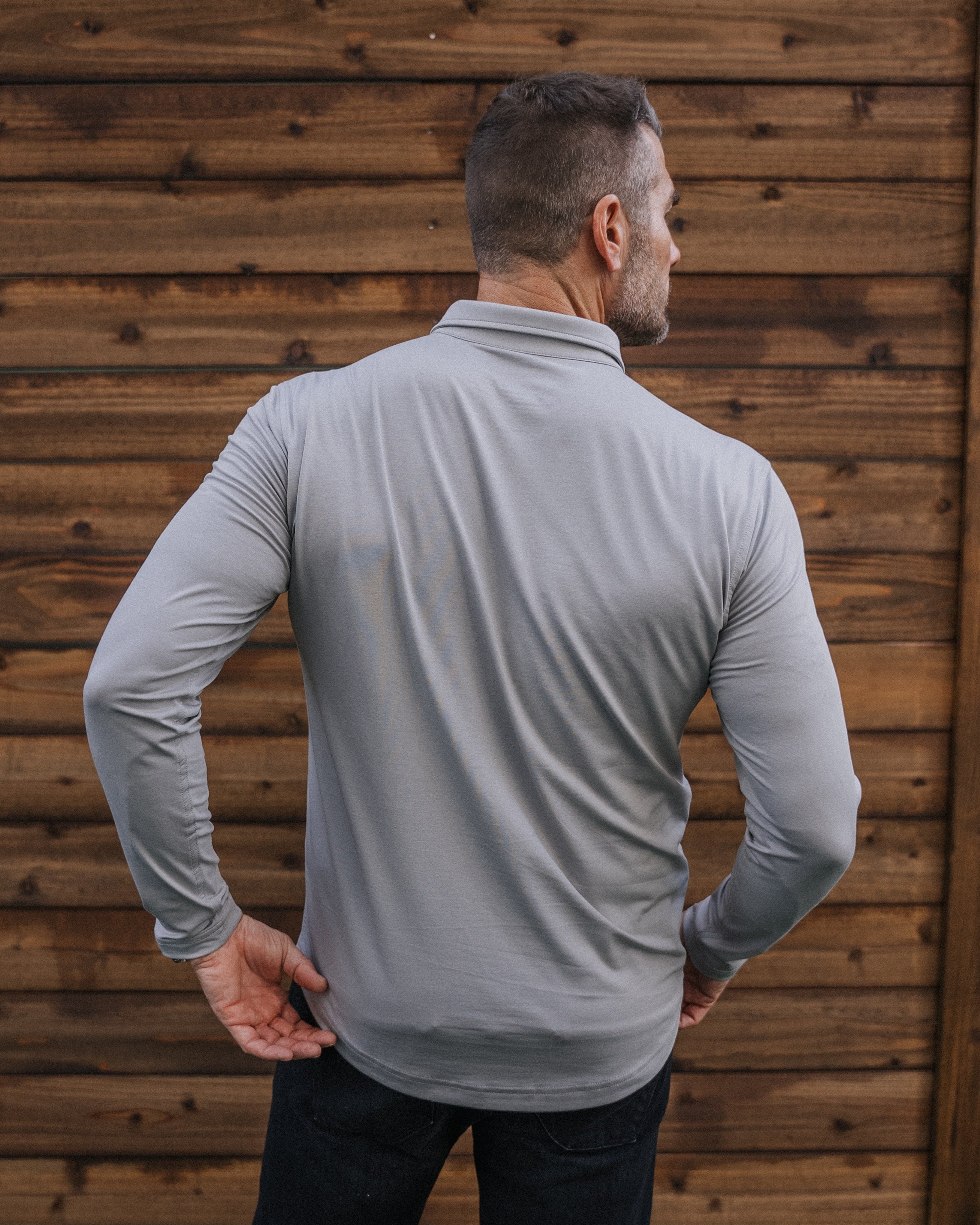 A man viewed from behind wearing a light gray long-sleeve quarter-zip performance shirt and dark pants, standing in front of a brown wooden fence.