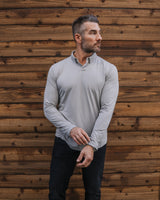 A man wearing a light gray long-sleeve quarter-zip pullover, black pants, and white sneakers stands in front of a wooden fence and green artificial turf.