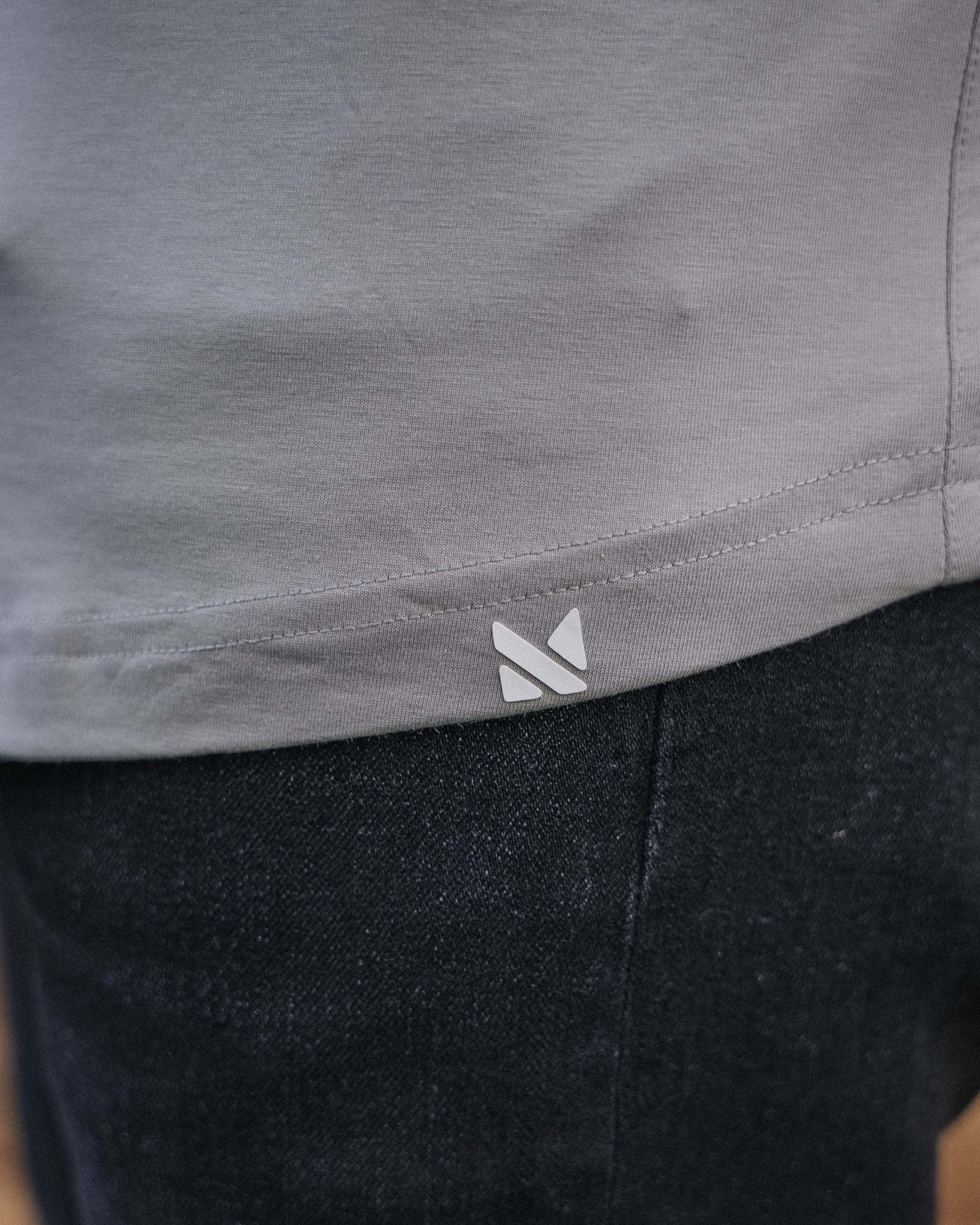 Close-up of a person wearing a light gray top and dark charcoal pants, with a small white geometric logo near the hem of the top. The background shows a blurred wooden floor.