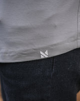 Close-up of a person wearing a light gray top and dark charcoal pants, with a small white geometric logo near the hem of the top. The background shows a blurred wooden floor.