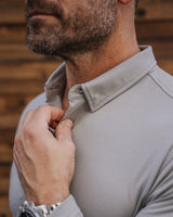 A close-up of a man wearing a light gray long-sleeve collared shirt, adjusting the collar with one hand while wearing a silver watch on the other wrist, set against a rustic wooden background.