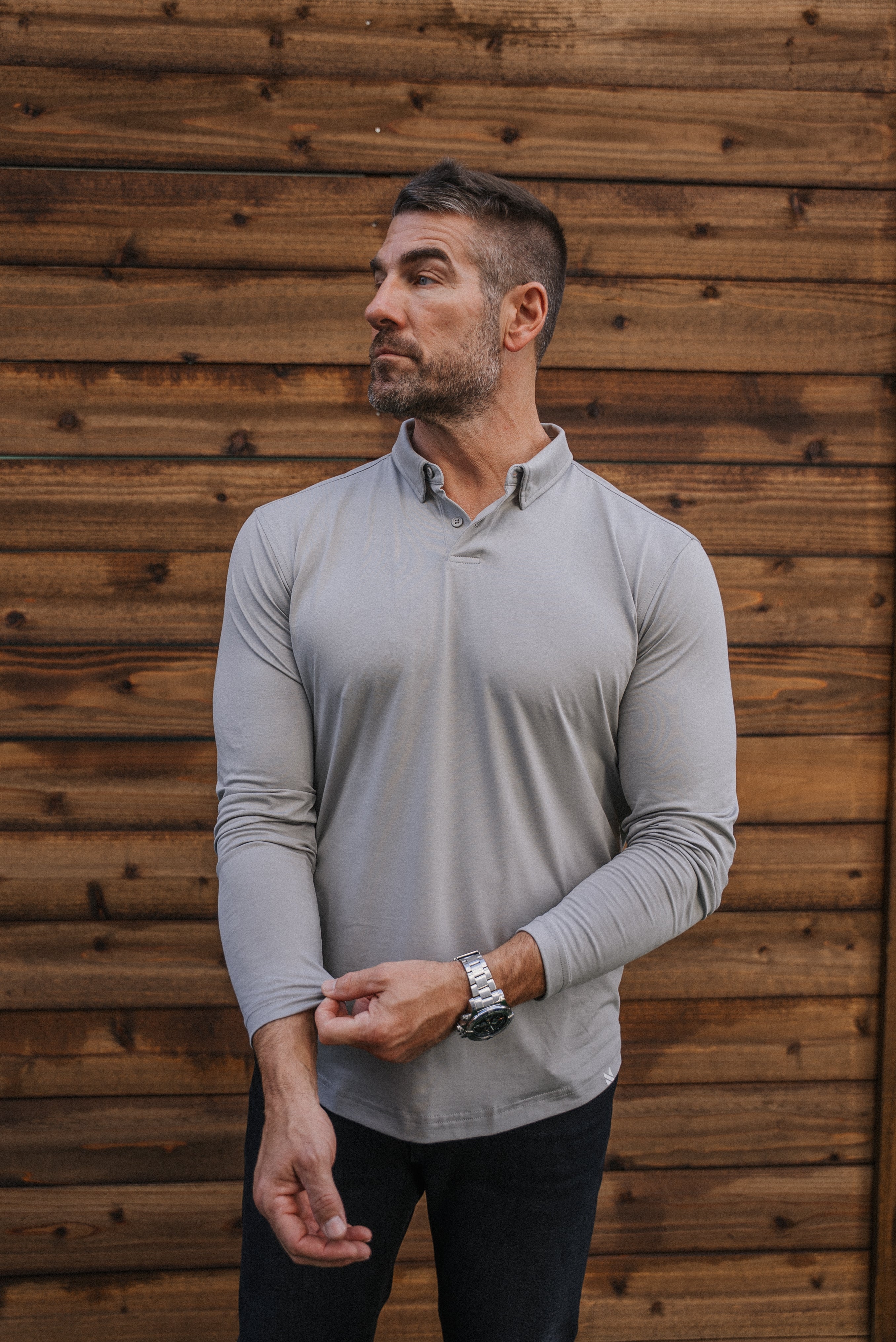 A man wearing a light gray long-sleeve collared athletic shirt and dark pants, standing in front of a rustic wooden plank wall.