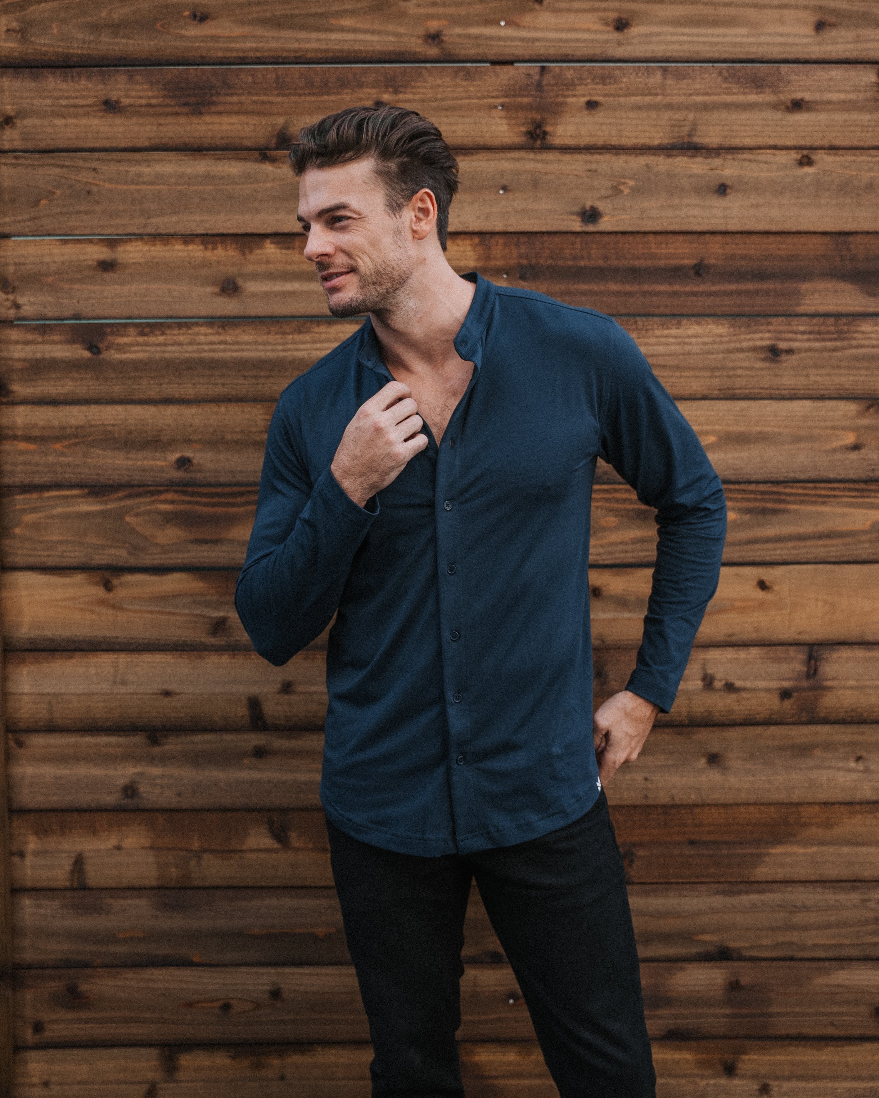 A man wearing a dark navy blue long-sleeve button-down shirt and black pants, standing against a rustic brown wooden plank wall.