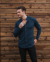 A man wearing a dark navy blue long-sleeve button-down shirt and black pants, standing against a rustic brown wooden plank wall.