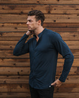 A man wearing a dark navy blue long-sleeve button-up shirt and black pants, standing against a rustic brown wooden plank wall.