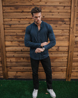 A man wearing a dark navy long-sleeve button-up shirt, black pants, and white sneakers stands on artificial green grass in front of a wooden fence, looking down at his wrist.