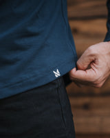 Close-up of a person wearing a dark navy blue t-shirt and black pants, pulling the hem of the shirt slightly to reveal a small white embroidered logo.