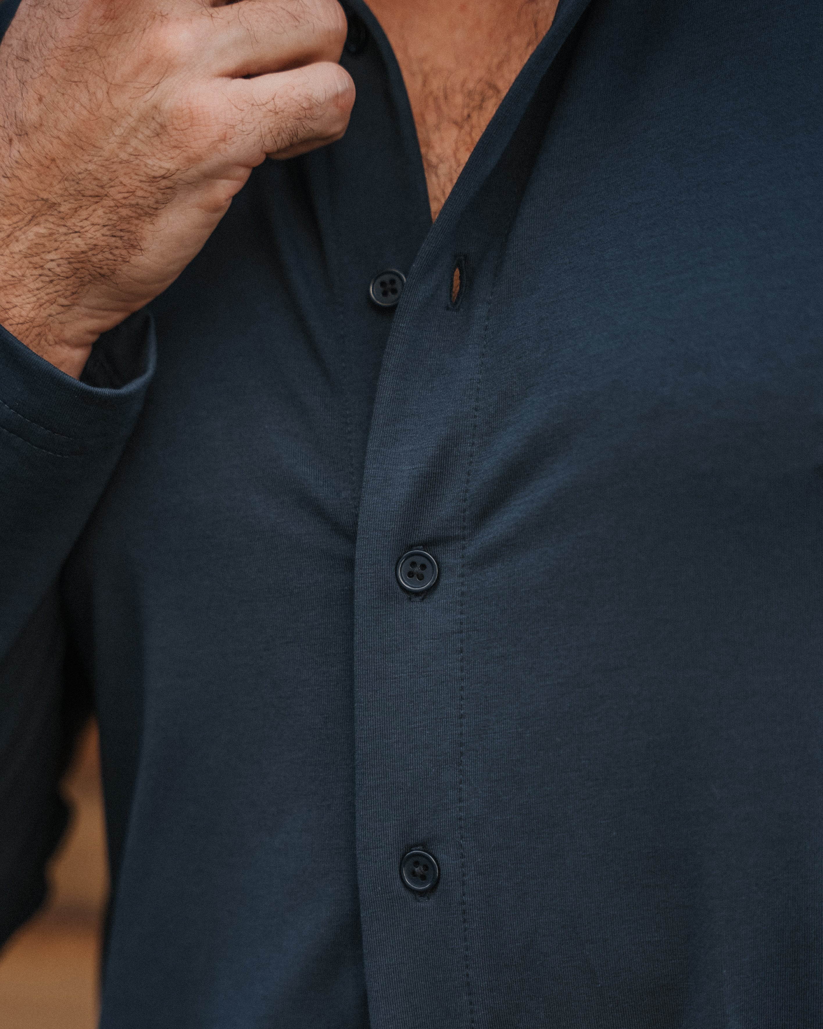 Close-up of a man wearing a dark navy long-sleeve button-up shirt, with his hands adjusting the collar and buttoning the front.