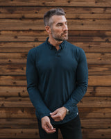 A man wearing a dark teal long-sleeve polo shirt and black pants, standing in front of a rustic brown wooden plank wall.