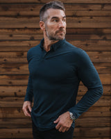 A man wearing a dark teal long-sleeve polo shirt and black pants, standing against a rustic brown wooden plank wall.