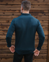 A man viewed from behind wearing a dark teal long-sleeve performance shirt and black pants, standing in front of a rustic brown wooden wall.