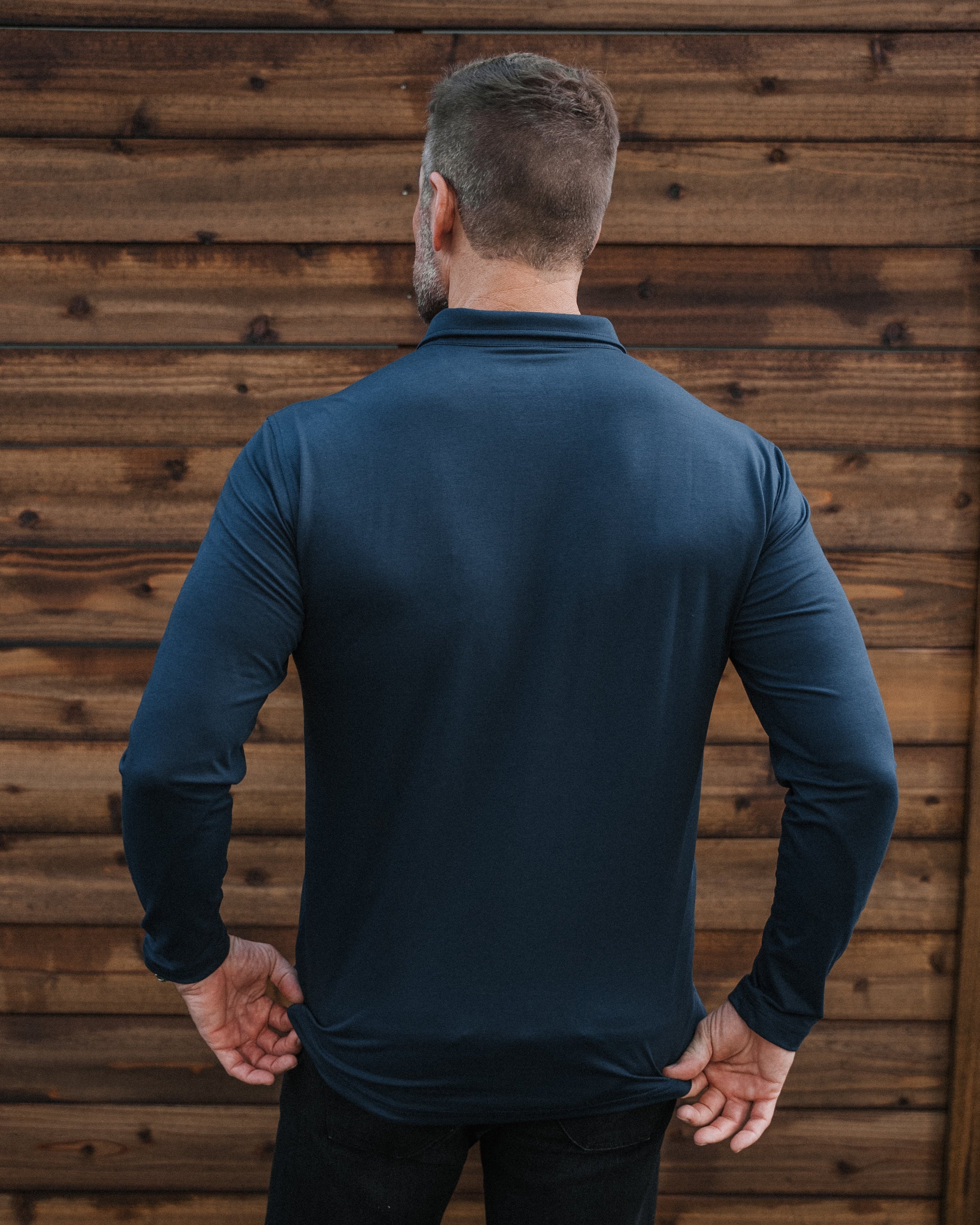 A man viewed from behind wearing a dark teal long-sleeve performance shirt and black pants, standing in front of a rustic brown wooden wall.