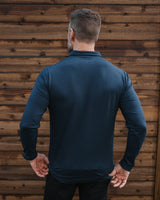 A man viewed from behind wearing a dark teal long-sleeve performance shirt and black pants, standing in front of a rustic brown wooden wall.