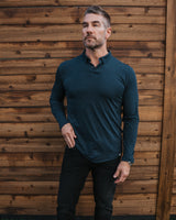A man wearing a dark teal long-sleeve polo shirt, black pants, and white sneakers stands in front of a wooden fence and artificial grass.