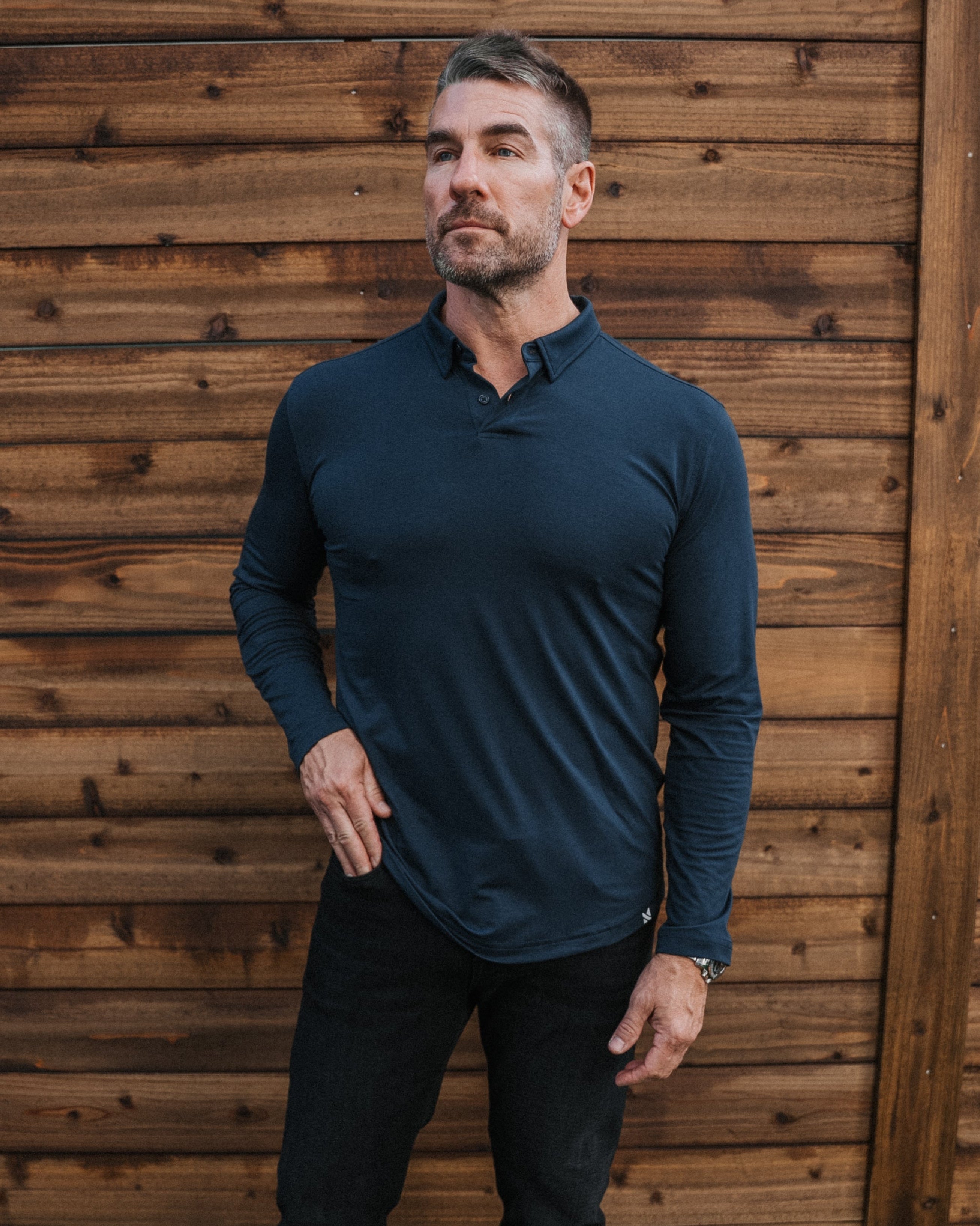 A man wearing a dark teal long-sleeve polo shirt, black pants, and white sneakers stands in front of a wooden fence and artificial grass.