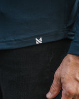 Close-up of a person wearing a dark navy long-sleeve top and black pants, with a small white geometric logo near the hem of the shirt and a hand resting near the waist.