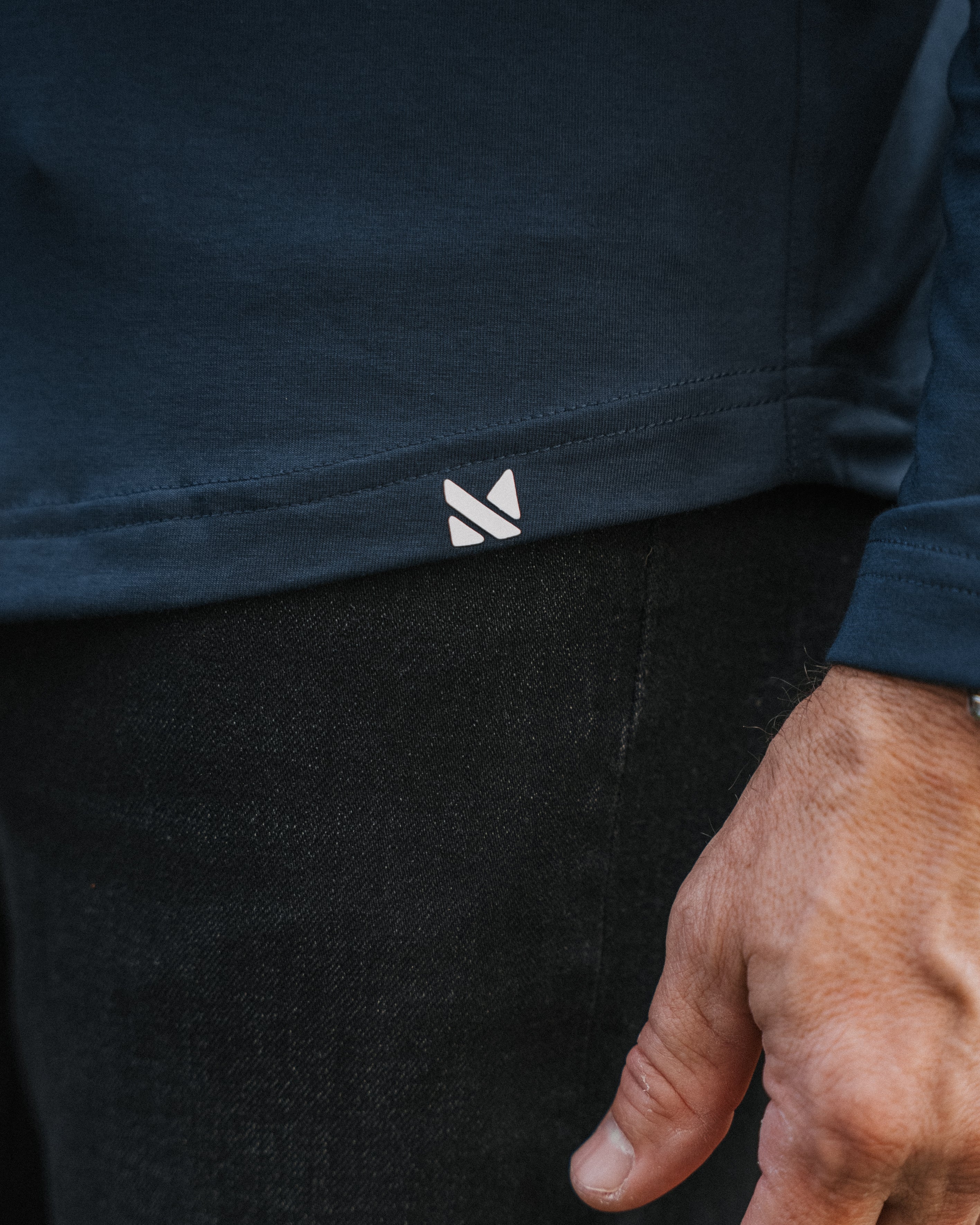 Close-up of a person wearing a dark navy long-sleeve top and black pants, with a small white geometric logo near the hem of the shirt and a hand resting near the waist.
