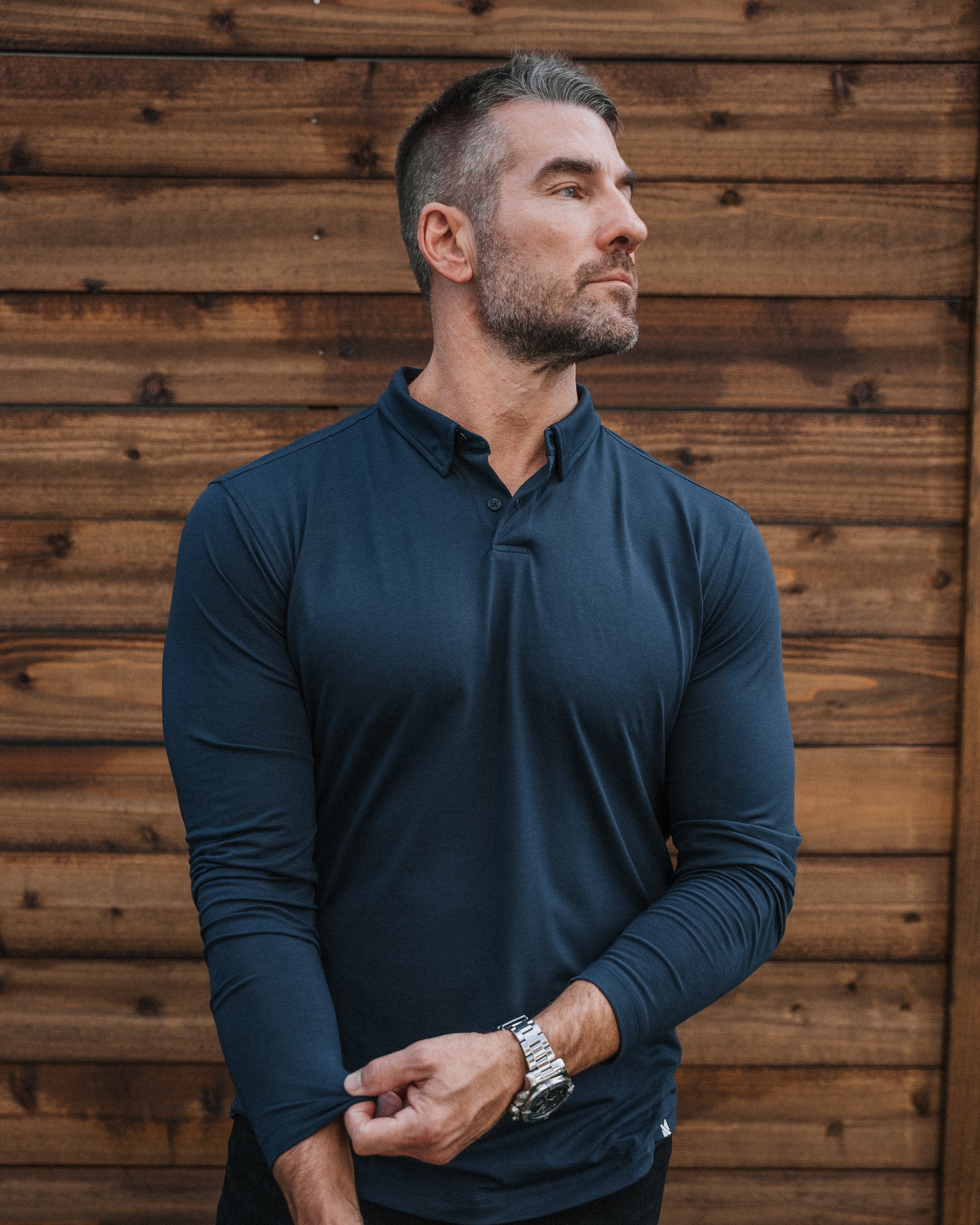 A man wearing a dark navy blue long-sleeve polo shirt with a buttoned collar, standing against a rustic brown wooden plank wall.