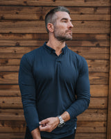 A man wearing a dark navy blue long-sleeve polo shirt with a buttoned collar, standing against a rustic brown wooden plank wall.