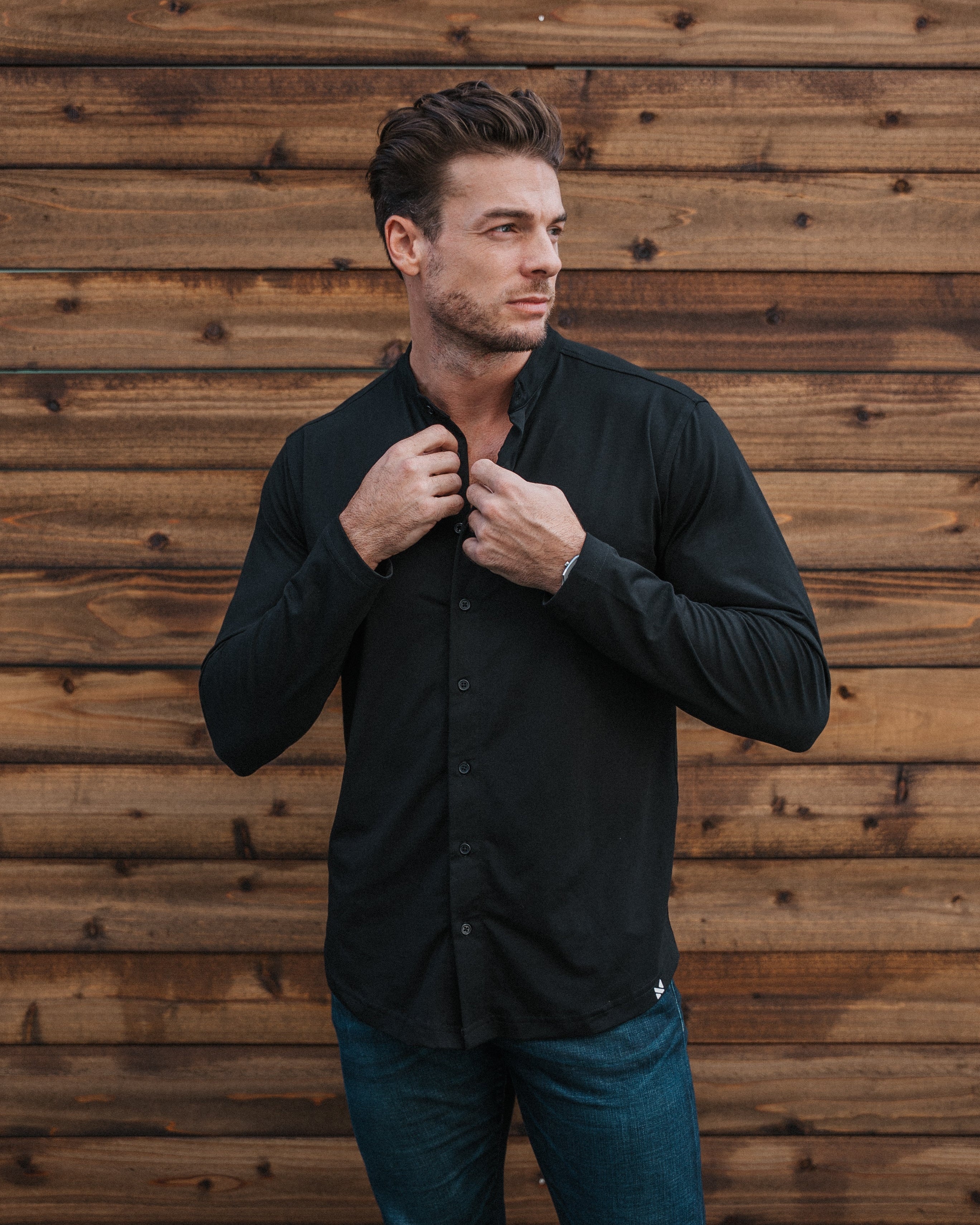 A man wearing a black long-sleeve button-up shirt and blue jeans, standing in front of a rustic brown wooden plank wall and adjusting the collar of his shirt.