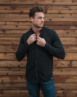 A man wearing a black long-sleeve button-up shirt and blue jeans, standing in front of a rustic brown wooden plank wall and adjusting the collar of his shirt.