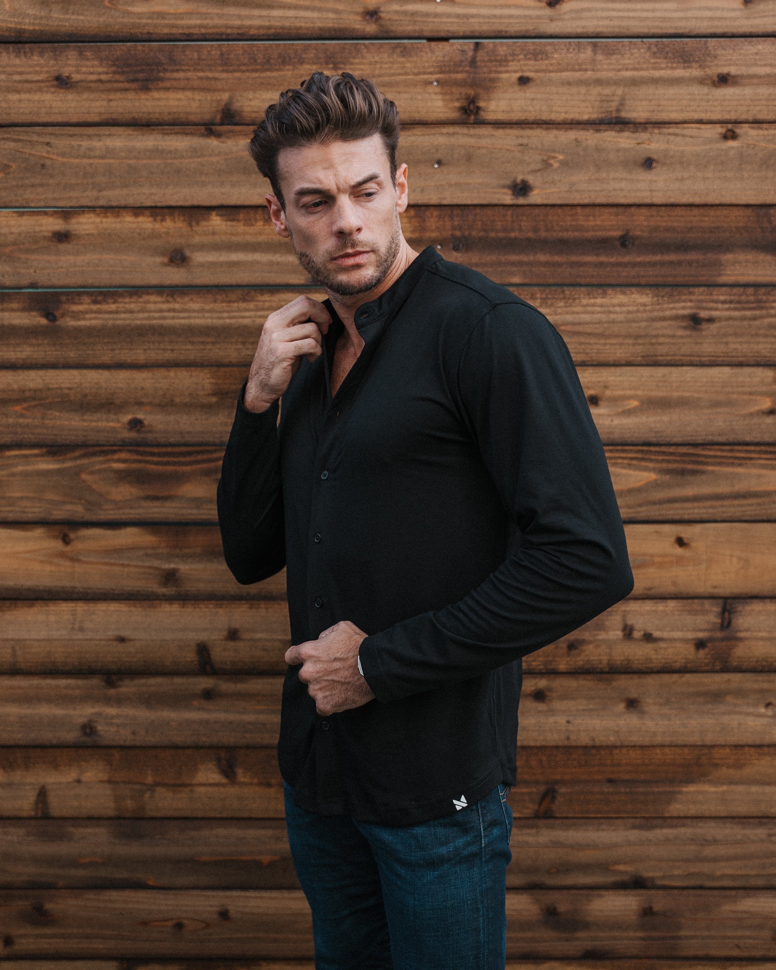 A man wearing a black long-sleeve button-up shirt and dark blue jeans, standing against a rustic brown wooden plank wall.