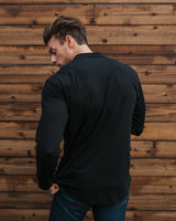 A man wearing a black long-sleeve crewneck shirt and blue jeans, photographed from behind against a rustic brown wooden plank wall.