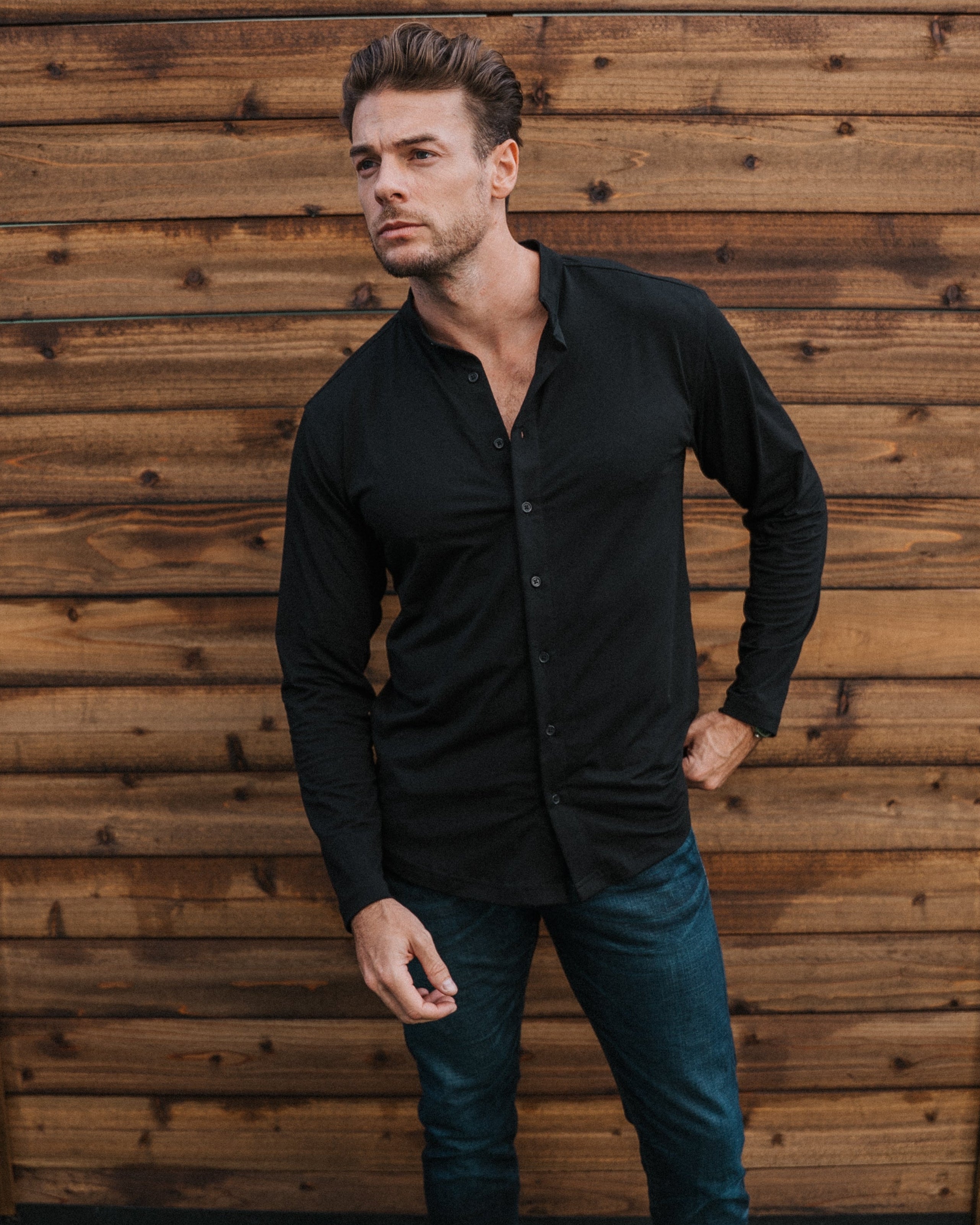 A man wearing a black long-sleeve button-up shirt, dark blue jeans, and white sneakers stands in front of a wooden fence and green artificial grass.