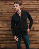 A man wearing a black long-sleeve button-up shirt, dark blue jeans, and white sneakers stands in front of a wooden fence and green artificial grass.