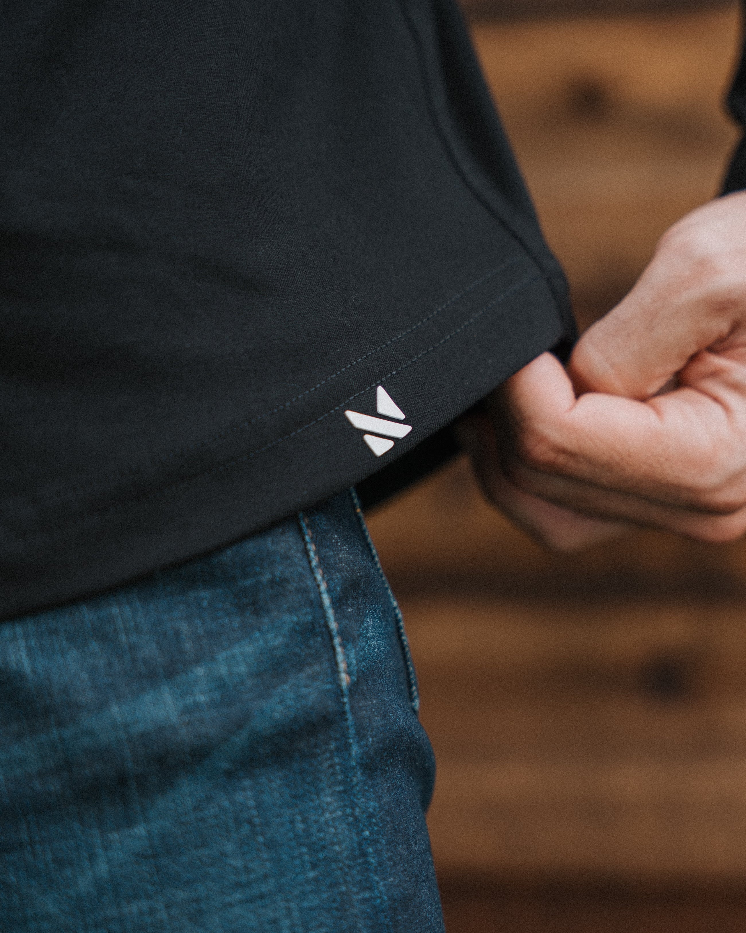 Close-up of a person wearing a black t-shirt and blue jeans, pulling the hem of the shirt down to reveal a small white geometric logo on the inside cuff. The background is a blurred wooden wall.