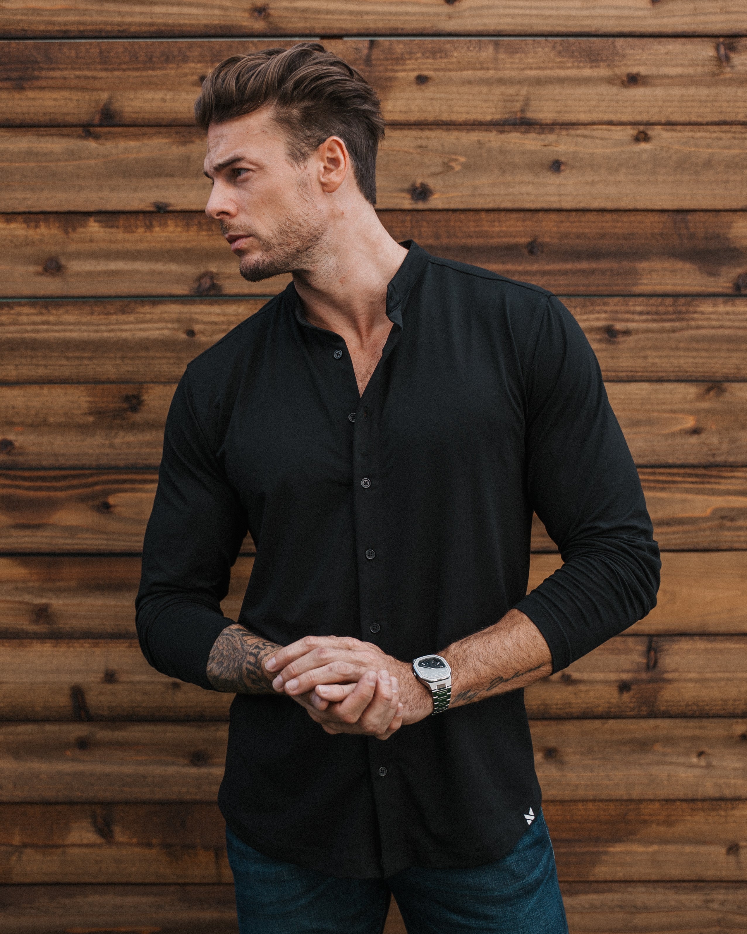 A man wearing a black long-sleeve button-up shirt and dark blue jeans, standing in front of a rustic wooden plank wall.