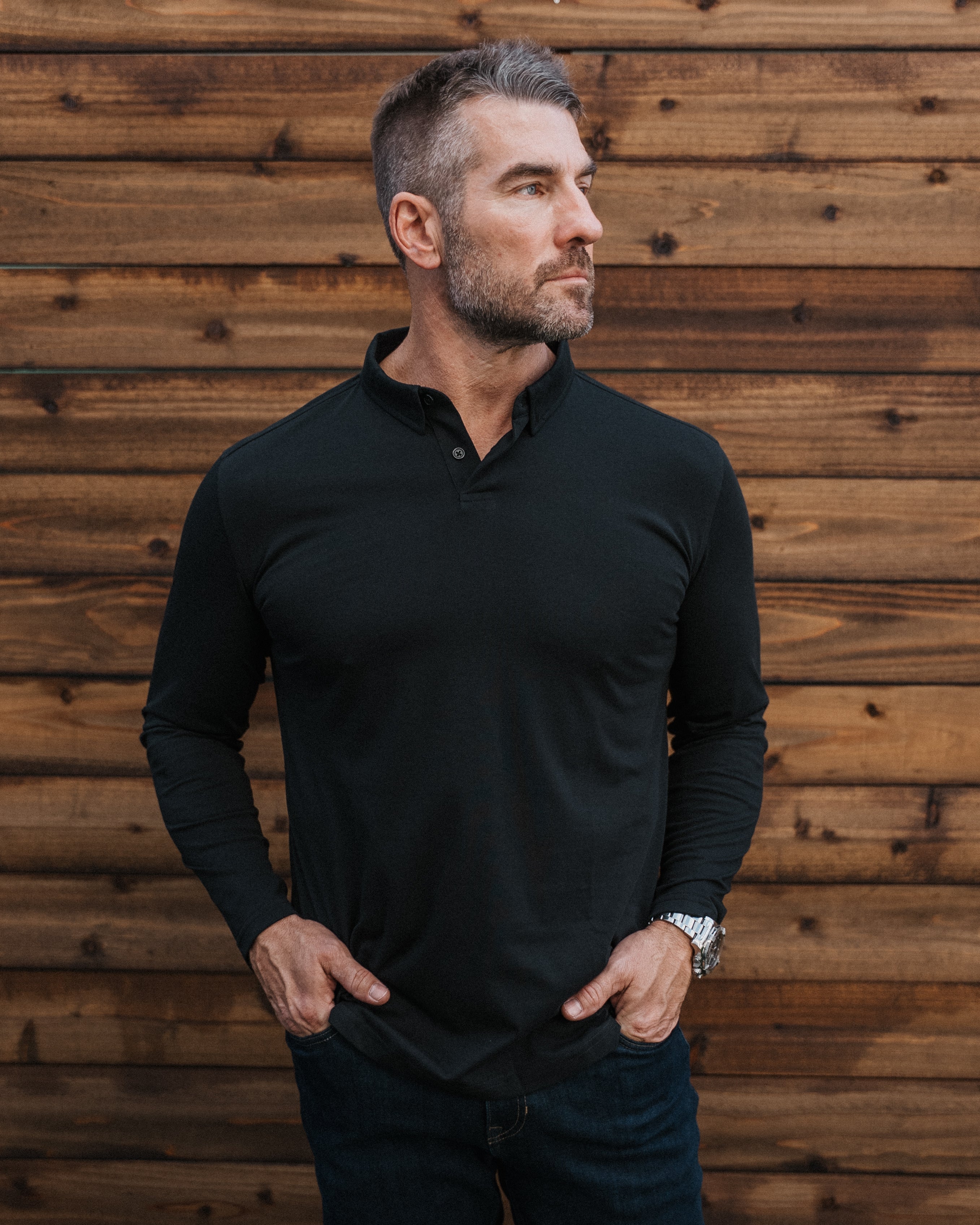 A man wearing a black long-sleeve polo shirt and dark jeans, standing against a rustic brown wooden plank wall.