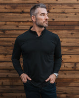 A man wearing a black long-sleeve polo shirt and dark jeans, standing against a rustic brown wooden plank wall.