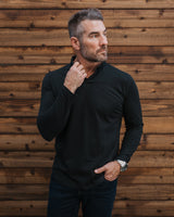 A man wearing a black long-sleeve polo shirt and dark jeans, standing against a rustic brown wooden plank wall.