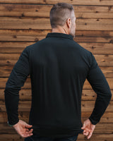 A man viewed from behind wearing a black long-sleeve collared shirt and dark jeans, standing in front of a rustic wooden plank wall.