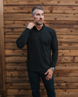 A man wearing a black long-sleeve polo shirt, dark jeans, and white sneakers stands in front of a wooden fence and artificial grass.