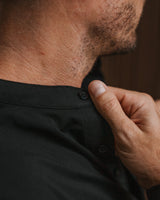 Close-up of a man wearing a black button-up shirt, adjusting the collar with his hand.
